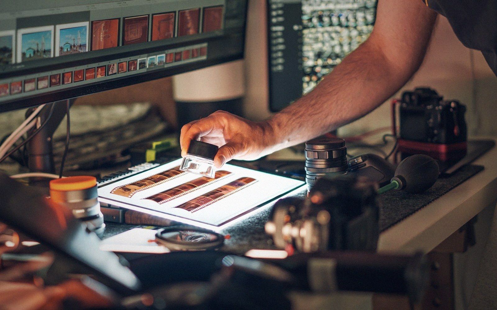A hand uses a loupe to examine film negatives on a lightbox beside a computer monitor